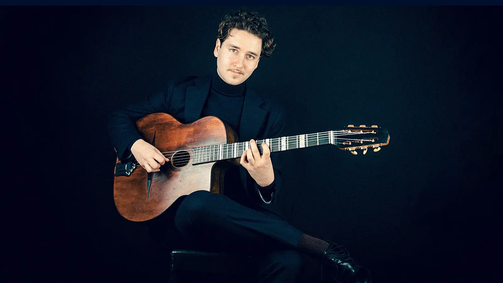 Porträtfoto von Elias Prinz mit Akustikgitarre, sitzend auf einem Hocker vor schwarzem Hintergrund, in dunklem Anzug und Rollkragenpullover während des Gitarrenspiels