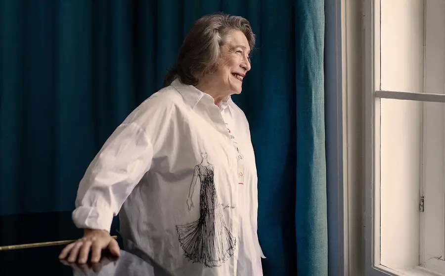 Elisabeth Leonskaja steht neben einem Fenster, trägt ein helles Hemd mit Zeichnung und blickt nach außen.