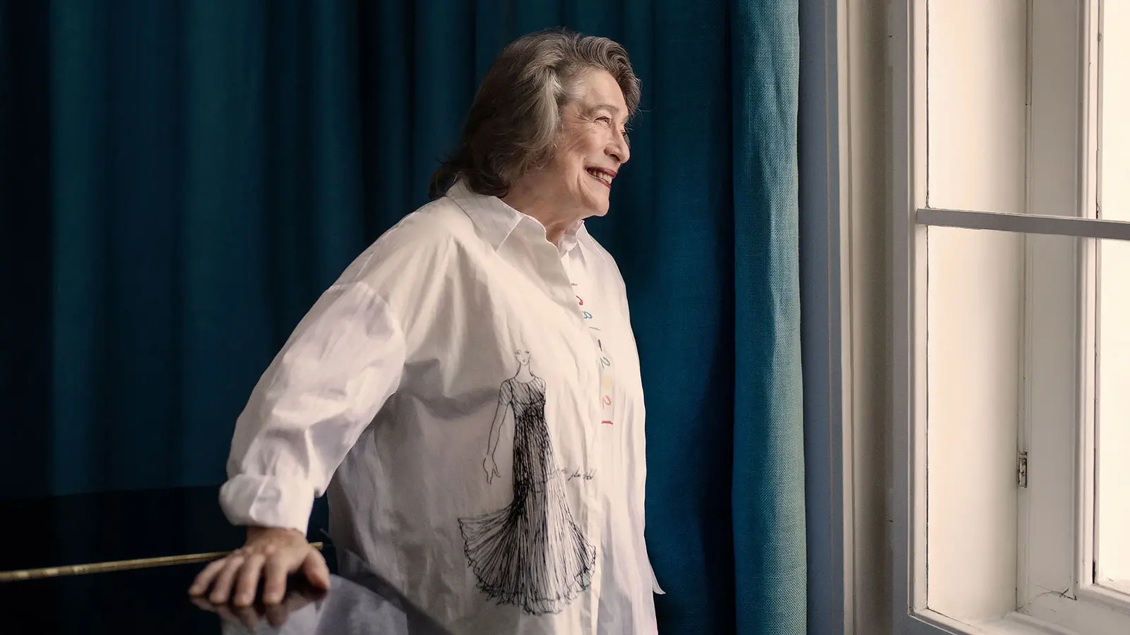 Elisabeth Leonskaja steht neben einem Fenster, trägt ein helles Hemd mit Zeichnung und blickt nach außen.