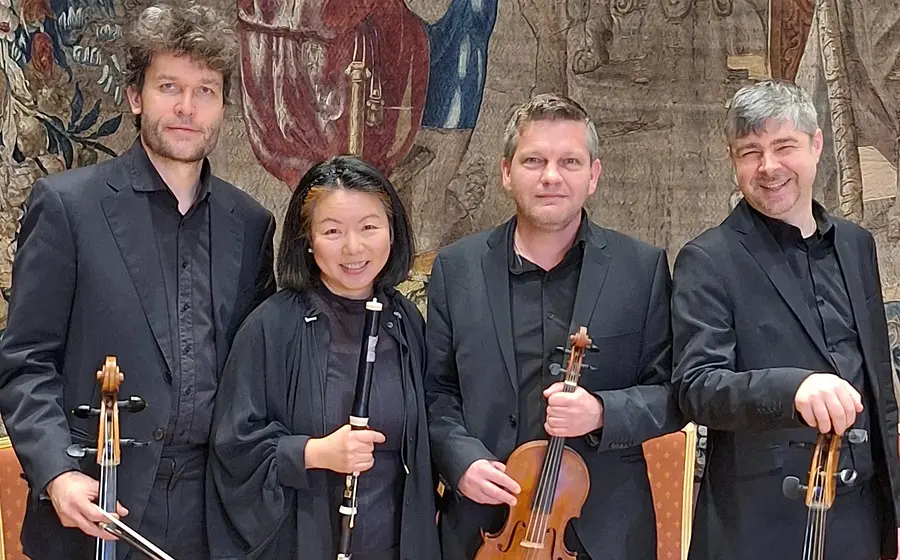 Vier Musikerinnen und Musiker in schwarzer Konzertkleidung stehen mit Celli, Klarinette und Violine vor einem historischen Wandteppich in einem Konzertsaal und blicken in die Kamera.