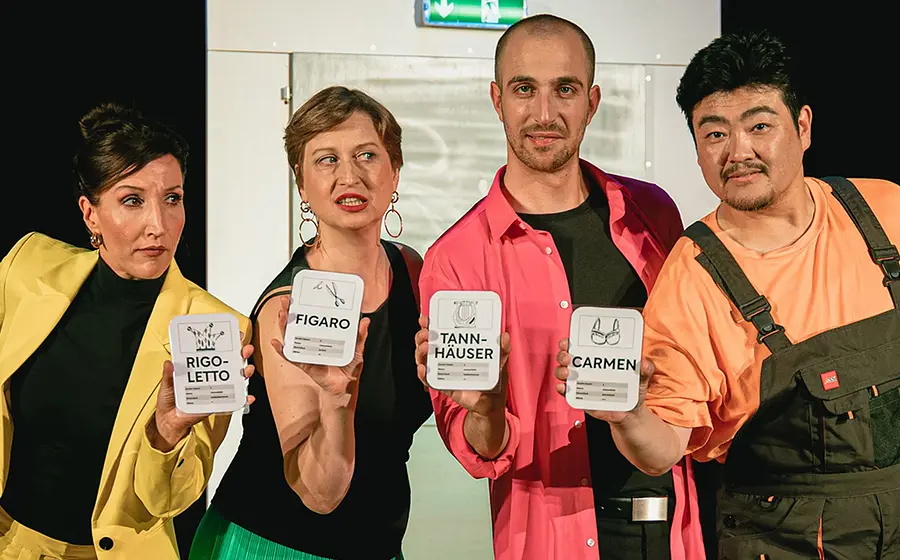 Bühnenfoto von Oper Plus mit vier Darstellenden vor heller Bühnenkulisse, die beschriftete Karten mit Operntiteln wie Rigoletto, Figaro, Tannhäuser und Carmen präsentieren, in farbenfroher Kleidung mit gelbem Blazer, pinkem Hemd und dunkler Latzhose.