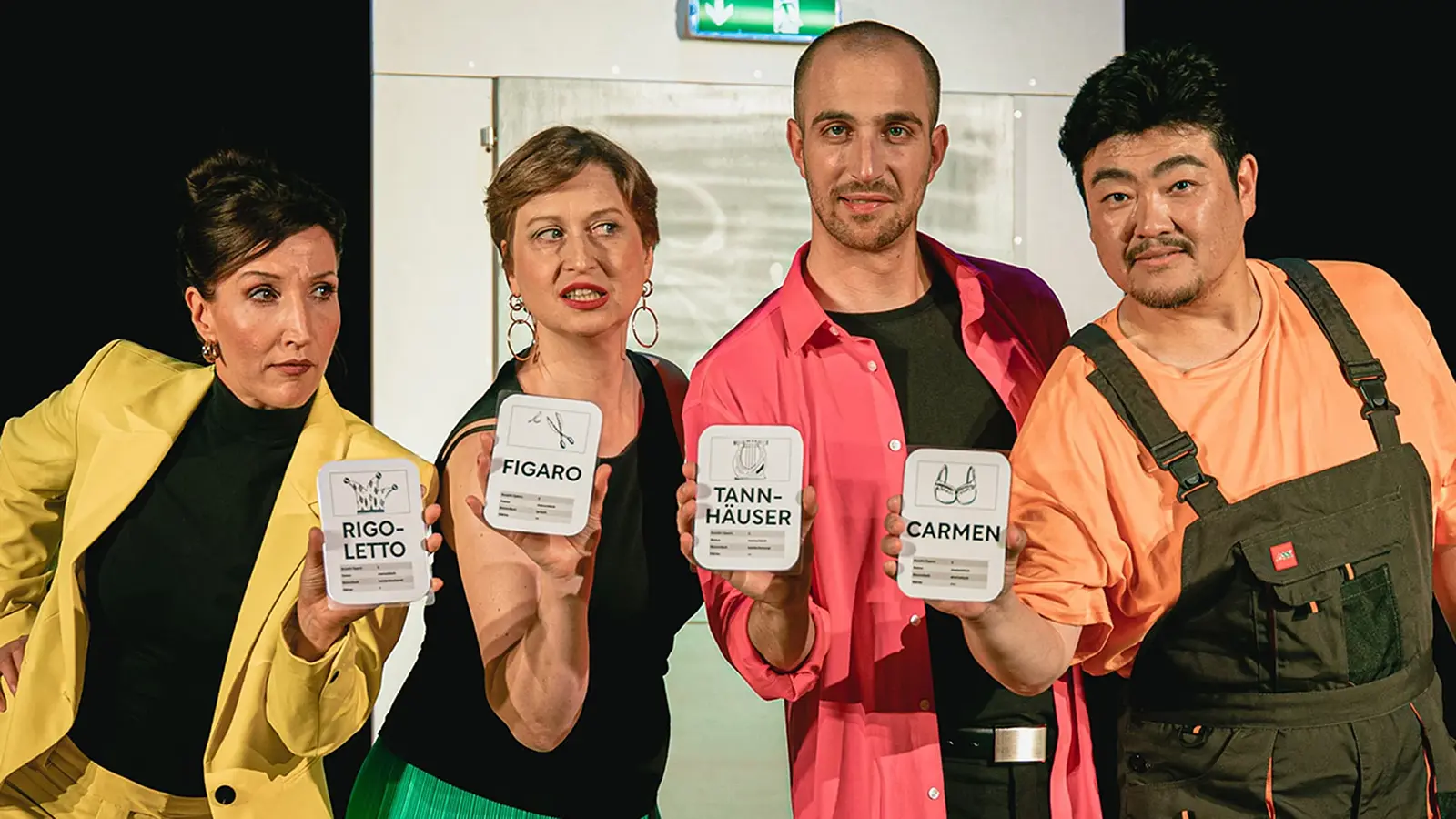 Bühnenfoto von Oper Plus mit vier Darstellenden vor heller Bühnenkulisse, die beschriftete Karten mit Operntiteln wie Rigoletto, Figaro, Tannhäuser und Carmen präsentieren, in farbenfroher Kleidung mit gelbem Blazer, pinkem Hemd und dunkler Latzhose.