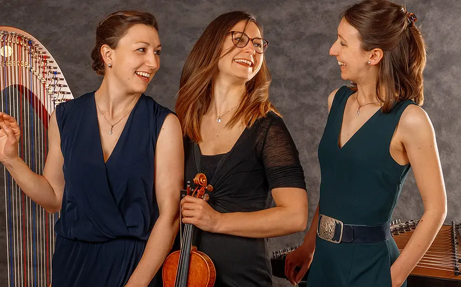 Foto für die Veranstaltung des Ensemble Vielsaitig mit den drei dunkel gekleidete, lachende Künstlerinnen mit Saiteninstrumenten und einer Harfe