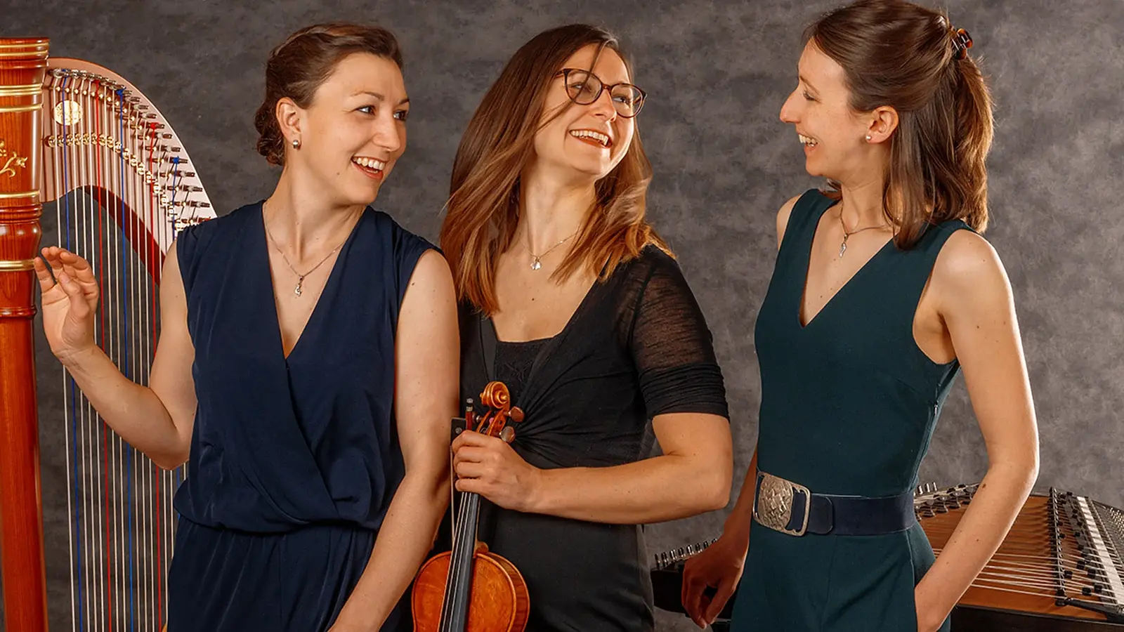 Foto für die Veranstaltung des Ensemble Vielsaitig mit den drei dunkel gekleidete, lachende Künstlerinnen mit Saiteninstrumenten und einer Harfe