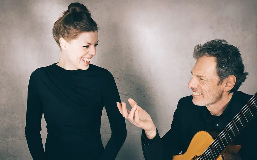 Zwei Personen vor grauer Wand, eine Person in schwarzer Kleidung, die zur anderen blickt, daneben eine Person mit klassischer Gitarre in schwarzem Sakko, beide einander zugewandt.