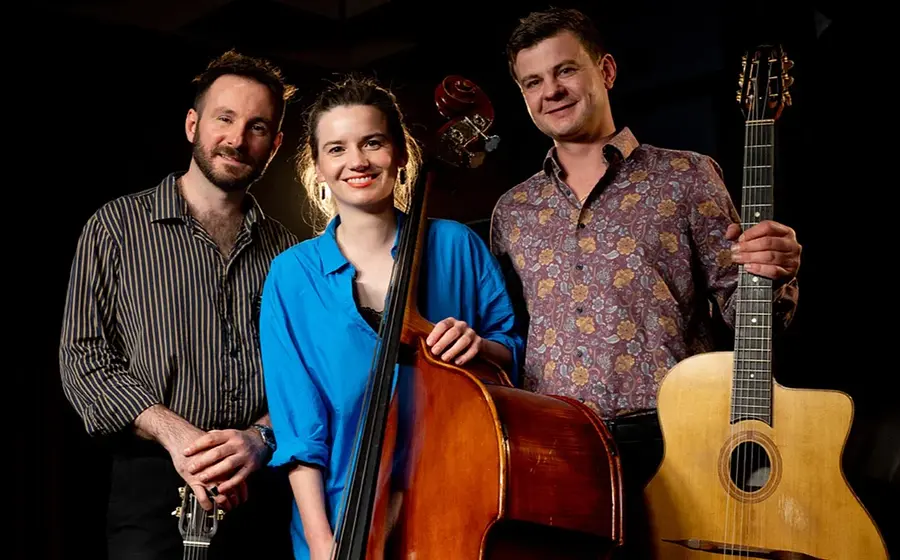 Die European Django Band mit drei Musikerinnen und Musikern vor dunklem Hintergrund, eine Person mit Kontrabass in blauem Hemd in der Mitte, flankiert von zwei Personen mit Gitarren.
