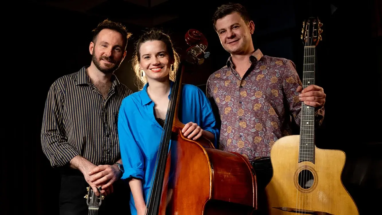 Die European Django Band mit drei Musikerinnen und Musikern vor dunklem Hintergrund, eine Person mit Kontrabass in blauem Hemd in der Mitte, flankiert von zwei Personen mit Gitarren.