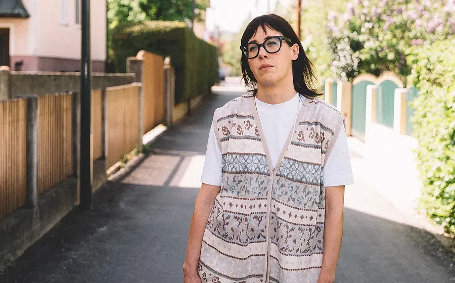 Eva Karl Faltermeier in einem weißen Shirt mit Weste steht in der Mitte eines Weges, links und rechts im Bild Gartenzäune und Sträucher sowie ein Haus