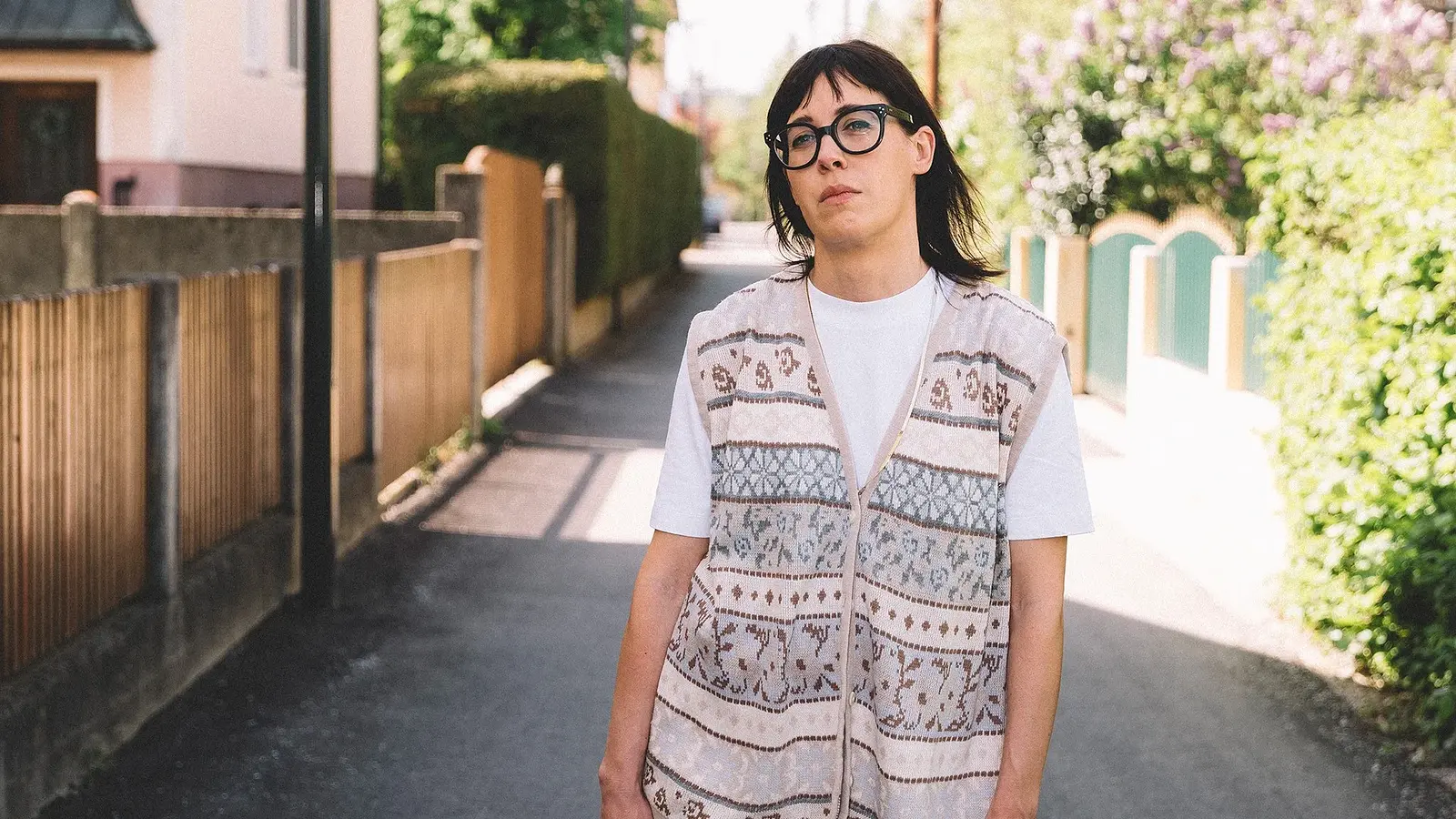 Eva Karl Faltermeier in einem weißen Shirt mit Weste steht in der Mitte eines Weges, links und rechts im Bild Gartenzäune und Sträucher sowie ein Haus