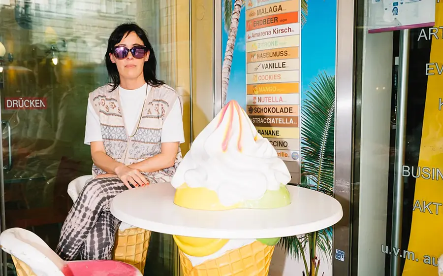Eva Karl Faltermeier mit Sonnenbrille sitzt an einem Tisch, der eine große Eiswaffel mit Eis darstellt, im Hintergrund ein Reiseplakat