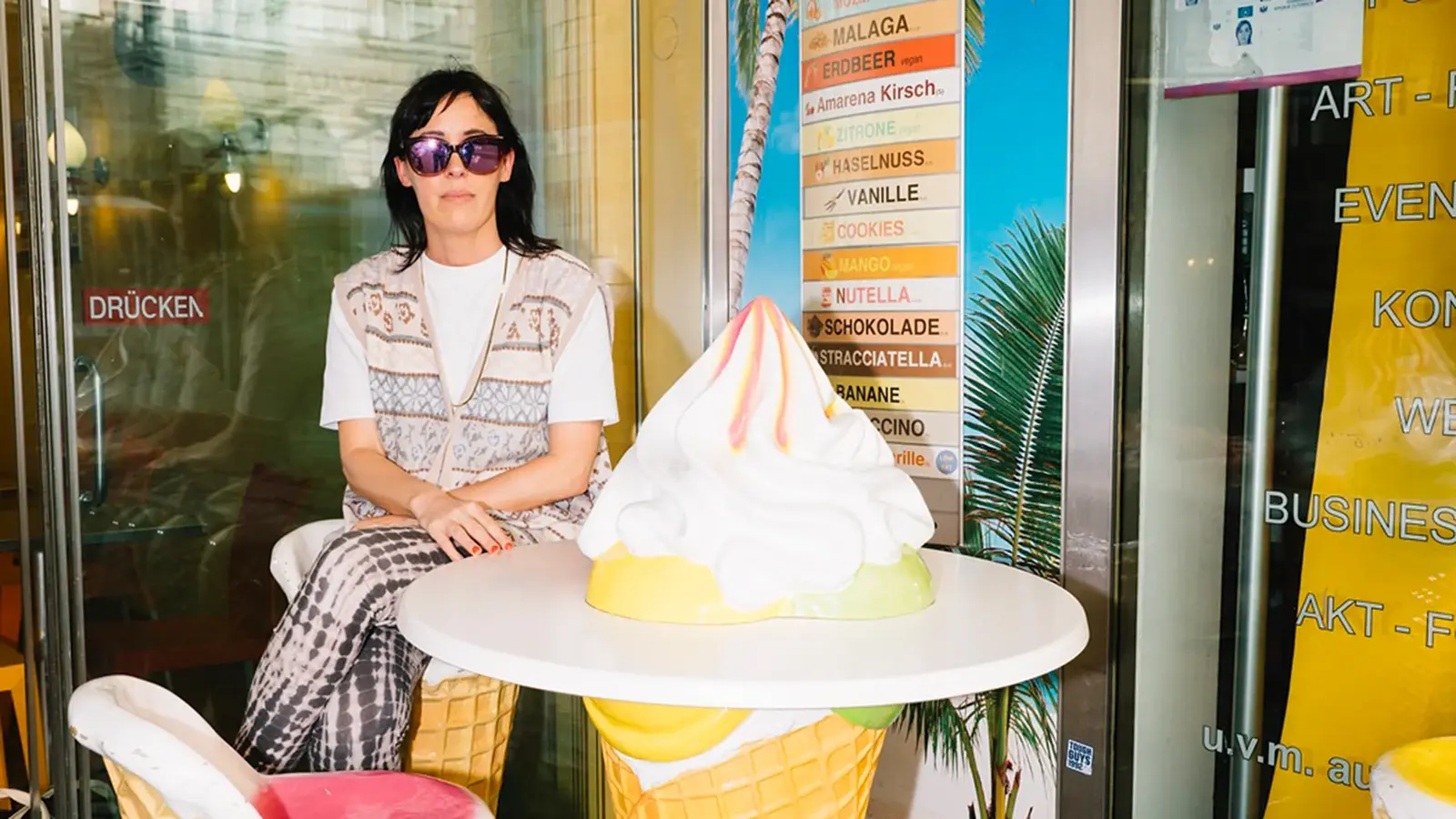 Eva Karl Faltermeier mit Sonnenbrille sitzt an einem Tisch, der eine große Eiswaffel mit Eis darstellt, im Hintergrund ein Reiseplakat