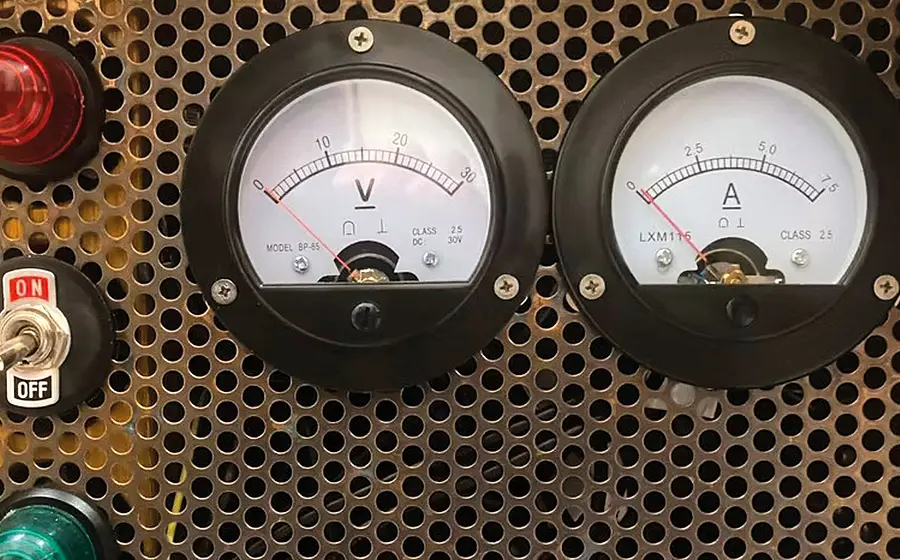 Nahaufnahme eines technischen Bedienfelds mit zwei analogen Messinstrumenten für Spannung und Stromstärke, einem roten und grünen Kontrolllicht sowie einem Kippschalter mit den Beschriftungen ON und OFF vor perforierter Metallfläche.