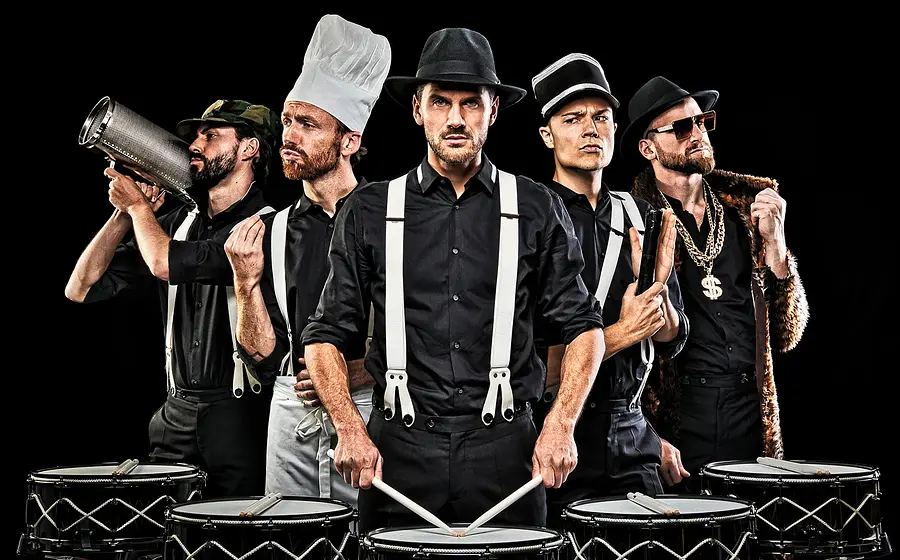 Fünf Performer in schwarzer Kleidung mit Hosenträgern stehen mit Trommeln und Percussion-Instrumenten vor schwarzem Hintergrund in unterschiedlichen Rollen