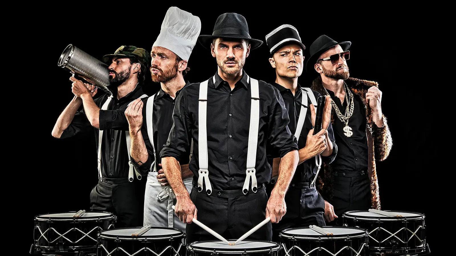 Fünf Performer in schwarzer Kleidung mit Hosenträgern stehen mit Trommeln und Percussion-Instrumenten vor schwarzem Hintergrund in unterschiedlichen Rollen