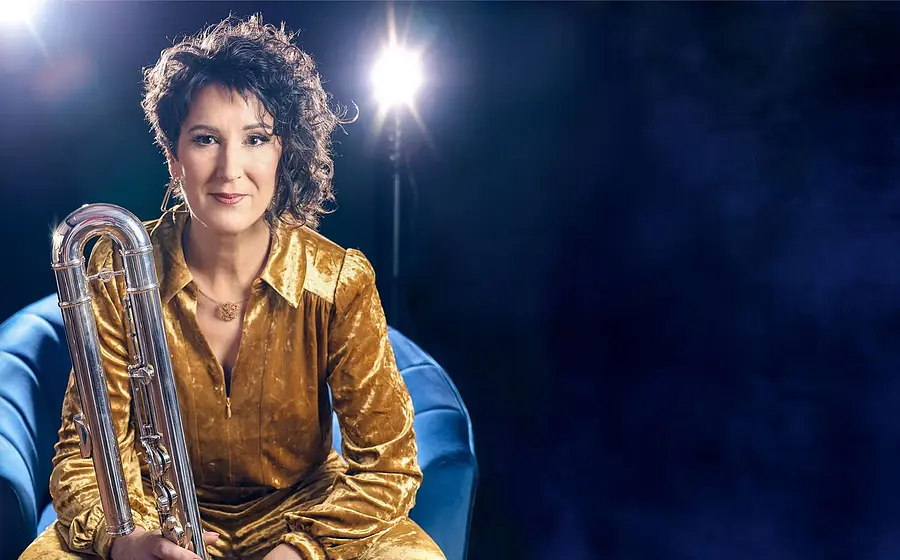 Isabelle Bodenseh auf einem blauen Sessel. Sie trägt goldfarbene Kleidung und hält eine silberne Bassklarinette. Dunkelblauer Hintergrund mit zwei hellen Scheinwerfern.