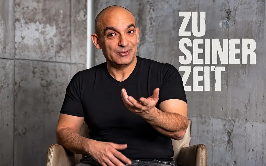 Porträt einer Person in schwarzem T Shirt auf einem Sessel vor grauer Betonwand. Die Person gestikuliert mit einer geöffneten Hand, daneben steht die Aufschrift Zu seiner Zeit.