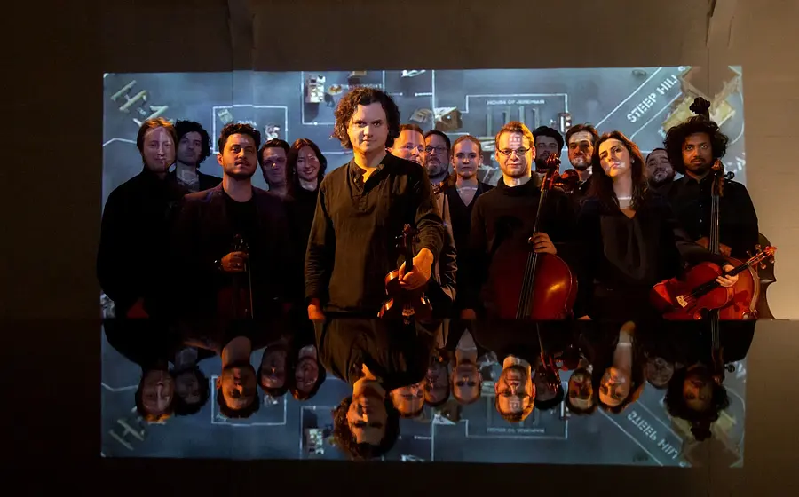 Mitglieder des dogma chamber orchestra mit Streichinstrumenten auf einer Bühne vor einer Videoprojektion.