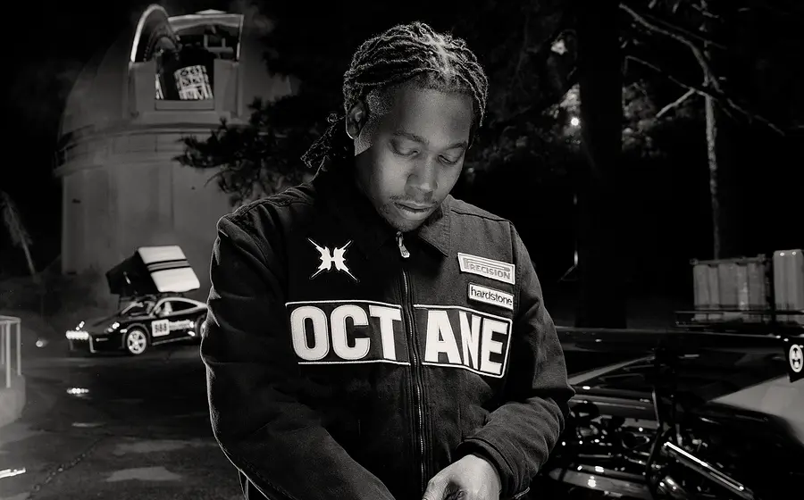 Schwarz‑weißes Foto von Don Toliver in „OCTANE“-Jacke, der nachts neben Autos und einem runden Gebäude nach unten schaut.