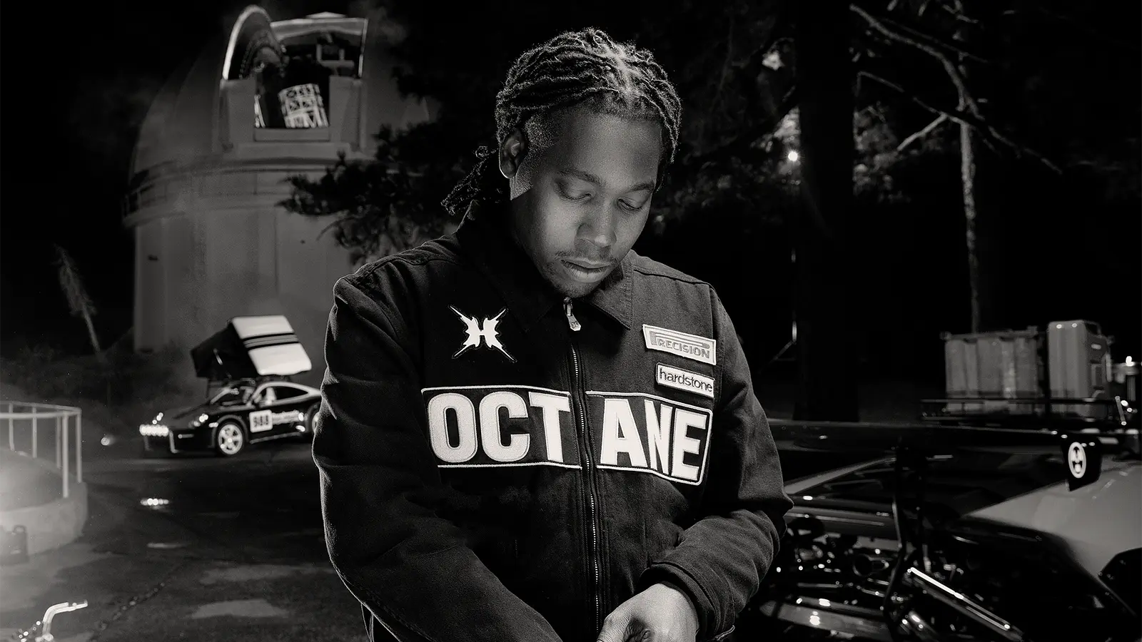 Schwarz‑weißes Foto von Don Toliver in „OCTANE“-Jacke, der nachts neben Autos und einem runden Gebäude nach unten schaut.
