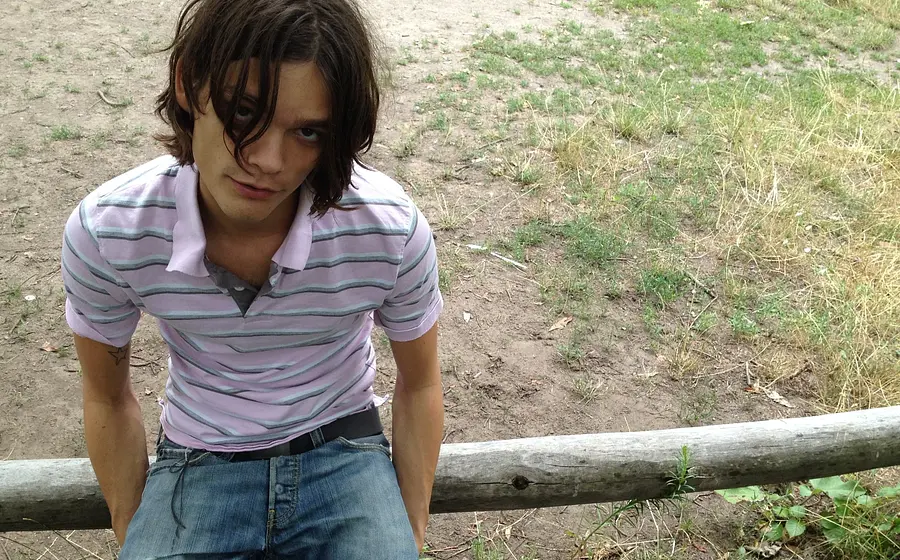 Porträtfoto von Donkey Kid in rosa gestreiftem Poloshirt und Jeans, auf einem Holzgeländer sitzend, mit schulterlangem Haar vor einer trockenen Wiesen- und Erdfläche im Freien.