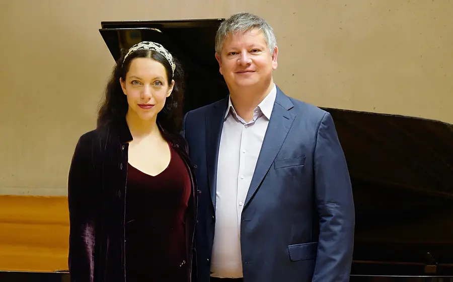 Thomas Groppner und Maharani Chakrabarti vor einem geöffneten Flügel, sie in dunkelrotem Samtkleid mit Haarband, er in blauem Anzug, vor hellem Konzertsaalhintergrund