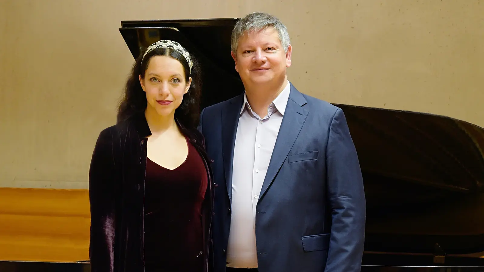 Thomas Groppner und Maharani Chakrabarti vor einem geöffneten Flügel, sie in dunkelrotem Samtkleid mit Haarband, er in blauem Anzug, vor hellem Konzertsaalhintergrund