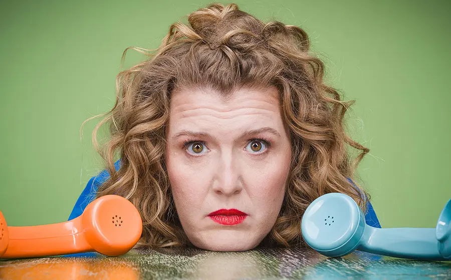 Franziska Wanninger stützt den Kopf auf eine Tischplatte zwischen einem blauen und einem orangen Retro-Telefonhörer, vor grünen Hintergrund