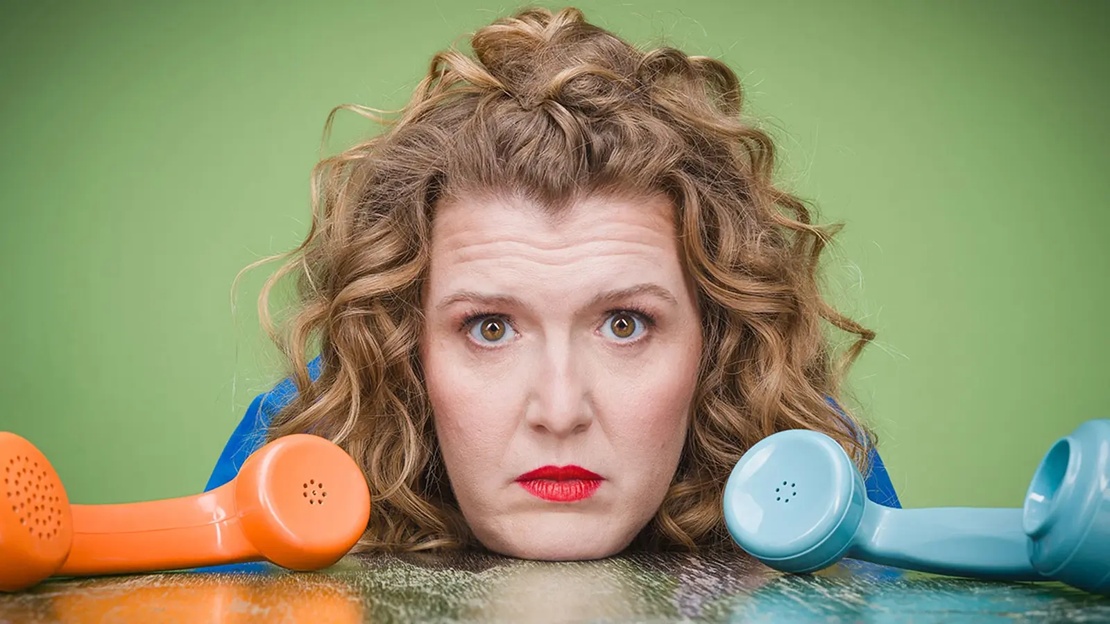 Franziska Wanninger stützt den Kopf auf eine Tischplatte zwischen einem blauen und einem orangen Retro-Telefonhörer, vor grünen Hintergrund