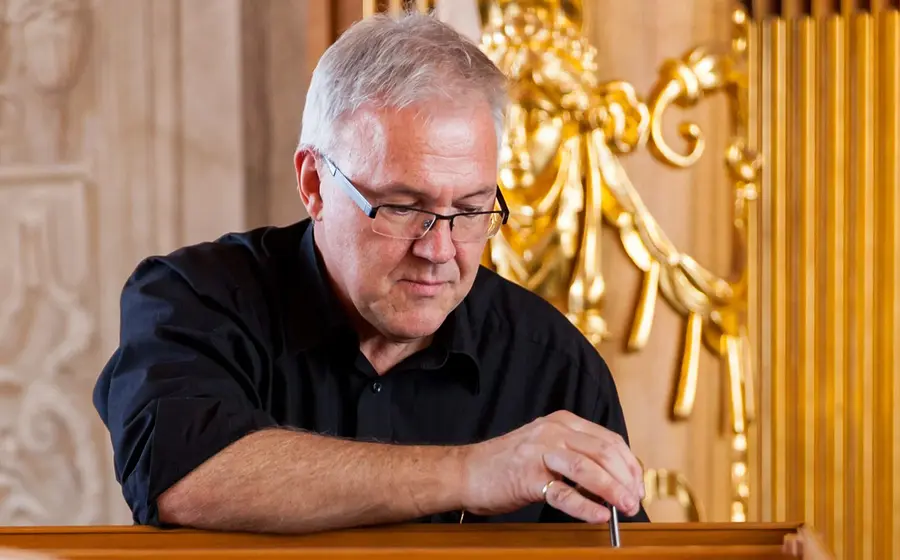 Person mit Brille an einer historischen Orgel in einem barocken Innenraum. Vergoldete Verzierungen und Orgelpfeifen prägen den Hintergrund, während eine Hand an den Bedienelementen des Instruments liegt