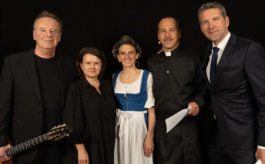Gruppenfoto mit fünf Personen vor schwarzem Hintergrund, darunter eine Person mit Gitarre, eine Person in schwarzer Kleidung, eine Person im blauen Dirndl, eine Person in schwarzer Priesterkleidung sowie eine Person im dunklen Anzug, nebeneinander für die Veranstaltung Frieden in Freiheit posierend.