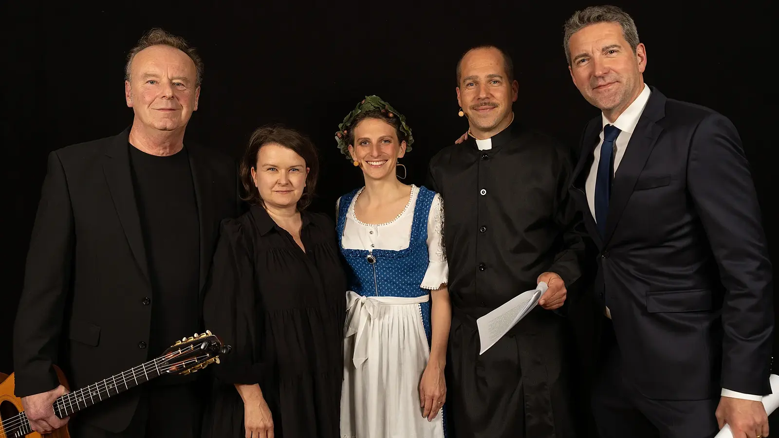 Gruppenfoto mit fünf Personen vor schwarzem Hintergrund, darunter eine Person mit Gitarre, eine Person in schwarzer Kleidung, eine Person im blauen Dirndl, eine Person in schwarzer Priesterkleidung sowie eine Person im dunklen Anzug, nebeneinander für die Veranstaltung Frieden in Freiheit posierend.