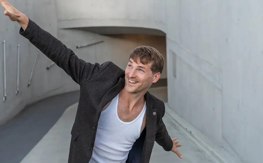 Person mit ausgestreckten Armen in einem geschwungenen Betonflur, aufgenommen aus erhöhter Perspektive vor moderner Architektur