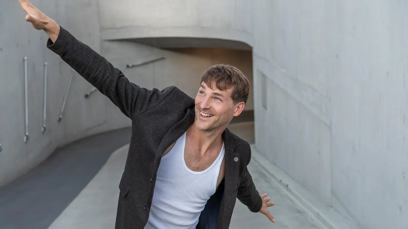 Person mit ausgestreckten Armen in einem geschwungenen Betonflur, aufgenommen aus erhöhter Perspektive vor moderner Architektur