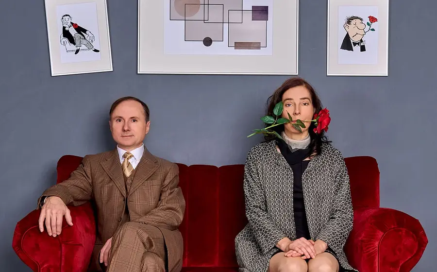 Zwei Personen sitzen auf einem roten Sofa vor grauer Wand mit gerahmten Illustrationen, eine Person im Anzug links, eine Person rechts mit roter Rose im Mund, beide frontal zur Kamera.
