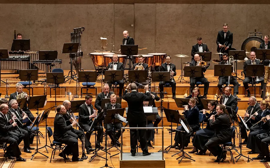 Großes Blasorchester in formaler Konzertkleidung für die Veranstaltung Gala del Centenario. Im Zentrum dirigiert eine Person das Ensemble, während Holzbläser, Blechbläser, Schlagwerk und Harfe in mehreren Reihen aufgestellt sind. Im Hintergrund sind Pauken, Gong und weiteres Schlagwerk zu sehen.