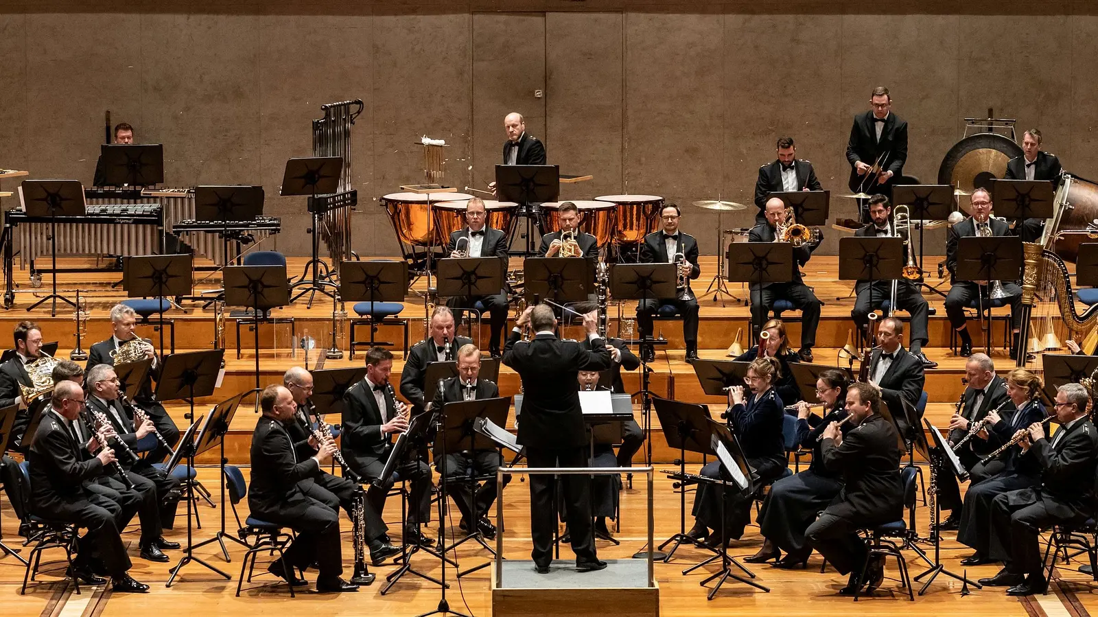 Großes Blasorchester in formaler Konzertkleidung für die Veranstaltung Gala del Centenario. Im Zentrum dirigiert eine Person das Ensemble, während Holzbläser, Blechbläser, Schlagwerk und Harfe in mehreren Reihen aufgestellt sind. Im Hintergrund sind Pauken, Gong und weiteres Schlagwerk zu sehen.