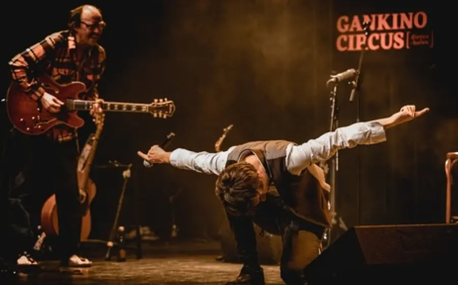 Bühnenfoto von Gankino Circus mit einem Musiker an der E‑Gitarre und einem weiteren Mitwirkenden mit ausgestreckten Armen und Mikrofon auf einer Konzertbühne, vor warmem Bühnenlicht und dem Schriftzug Gankino Circus im Hintergrund.
