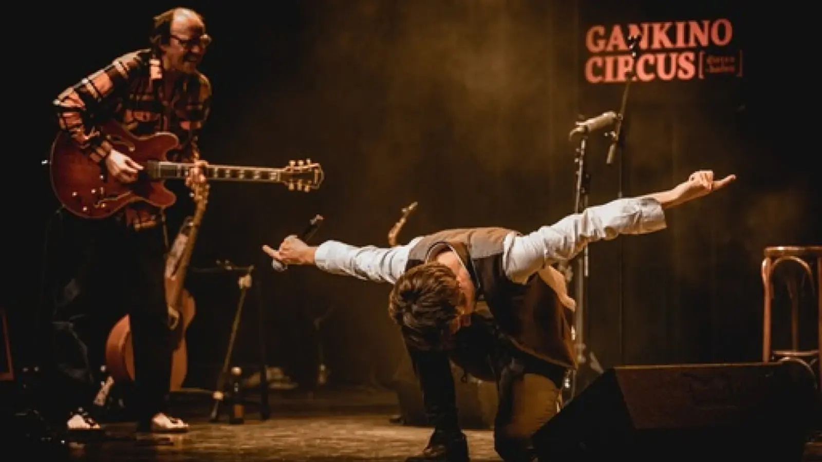 Bühnenfoto von Gankino Circus mit einem Musiker an der E‑Gitarre und einem weiteren Mitwirkenden mit ausgestreckten Armen und Mikrofon auf einer Konzertbühne, vor warmem Bühnenlicht und dem Schriftzug Gankino Circus im Hintergrund.