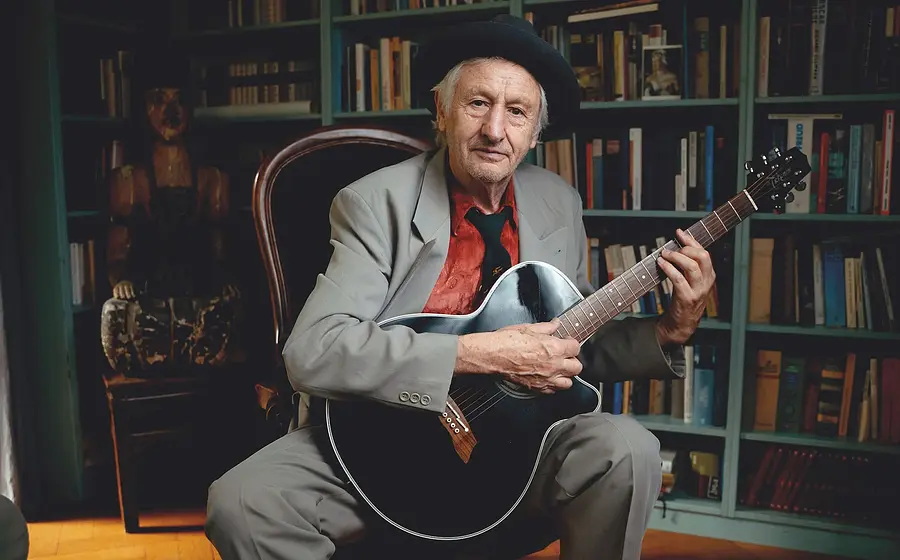 Foto für die Veranstaltung „Georg Ringsgwandl“ mit dem Künstler, der mit Hut auf einem Stuhl sitzt und eine Gitarre in der Hand hält.