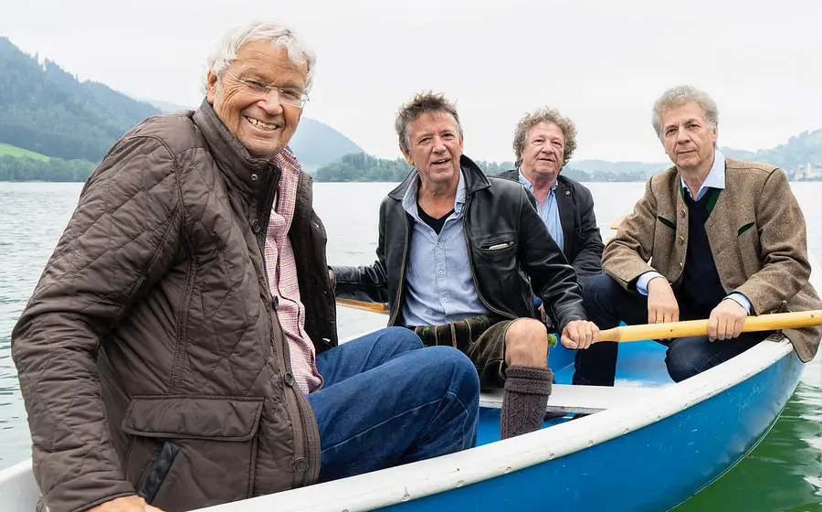 Gerhard Polt und die Well-Brüder auf einem bla-weißen Ruderboot auf einem See, im Hintergrund Berglandschaft