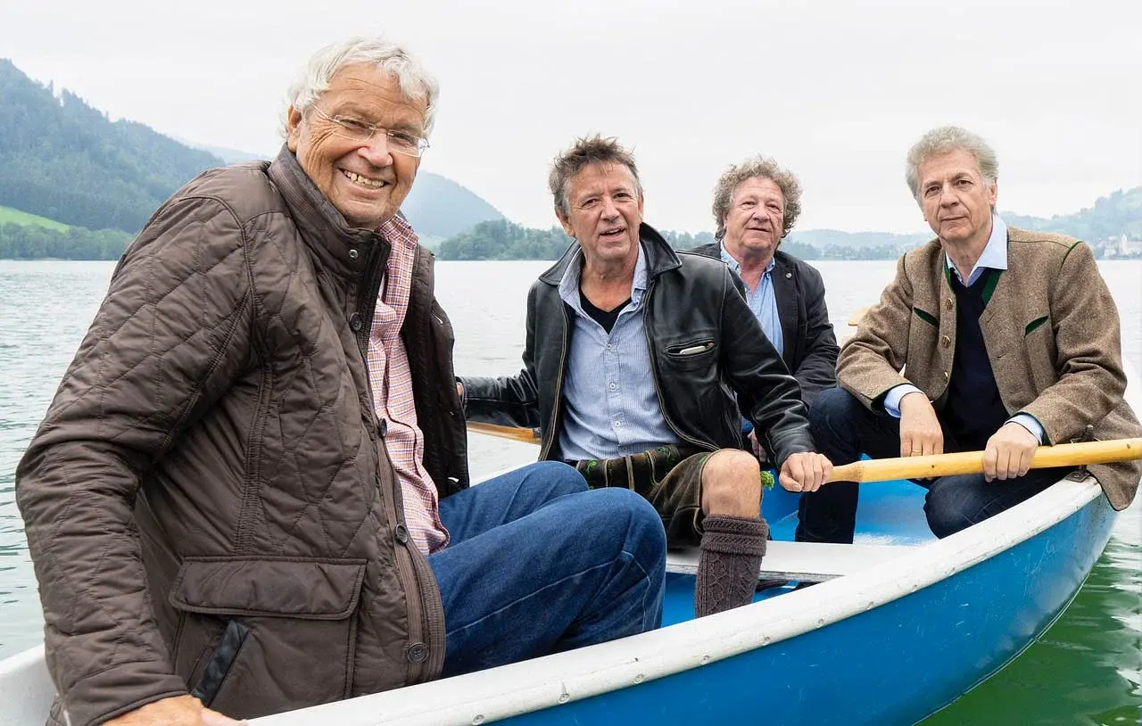 Gerhard Polt und die Well-Brüder auf einem bla-weißen Ruderboot auf einem See, im Hintergrund Berglandschaft