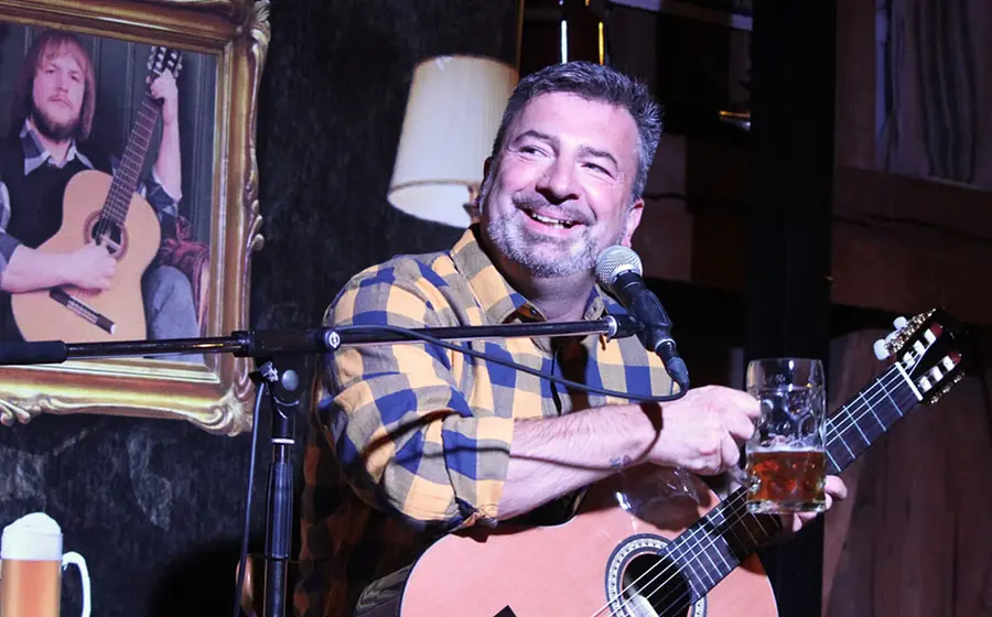 Foto zum Event von Gery Gerspitzer: Der Künstler mit Gitarre und Maßkrug, im Hintergrund ein Bild eines Mannes mit Gitarre