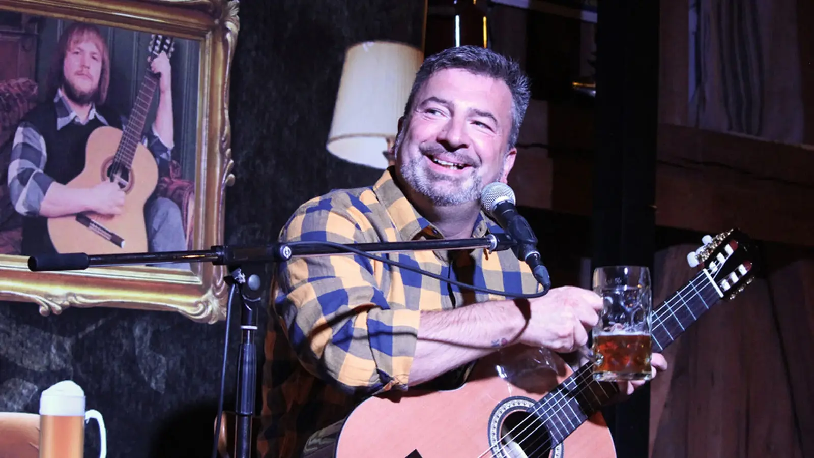 Foto zum Event von Gery Gerspitzer: Der Künstler mit Gitarre und Maßkrug, im Hintergrund ein Bild eines Mannes mit Gitarre