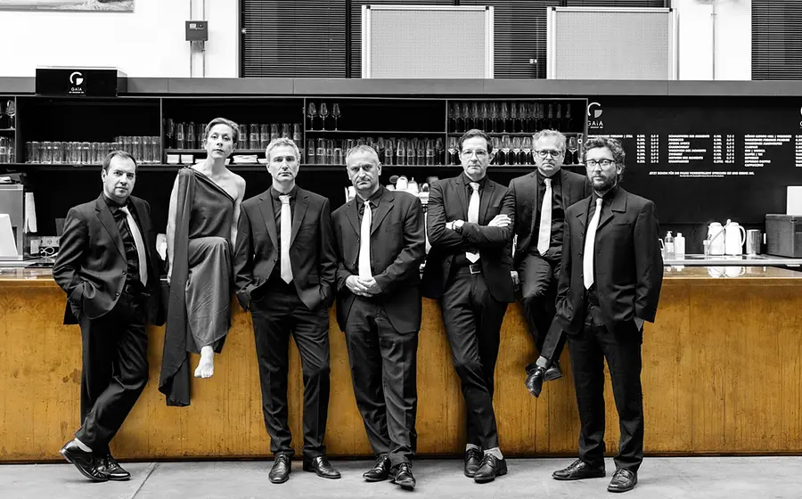 Bandfoto von Goldmund mit sieben Personen in dunkler Konzertkleidung vor einer Bar, sechs in Anzügen mit hellen Krawatten und eine Person in langem asymmetrischem Kleid, vor Regalen mit Gläsern und Getränkekarte im Hintergrund