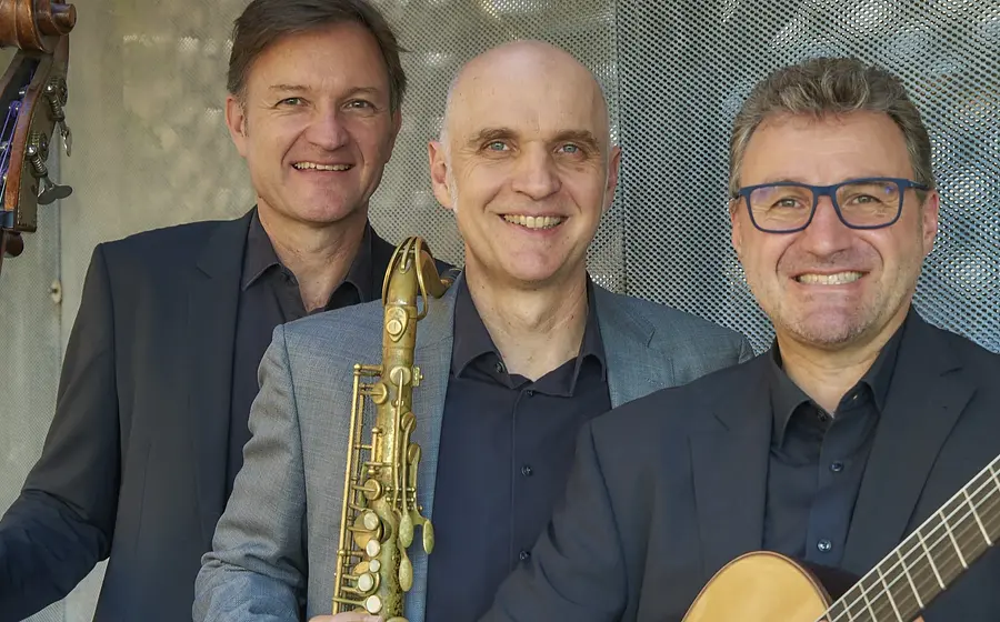 Die drei Mitglieder der Golden Notes in Anzügen mit Instrumenten lächeln in die Kamera, vor verschwommenen grau-blauem Hintergrund