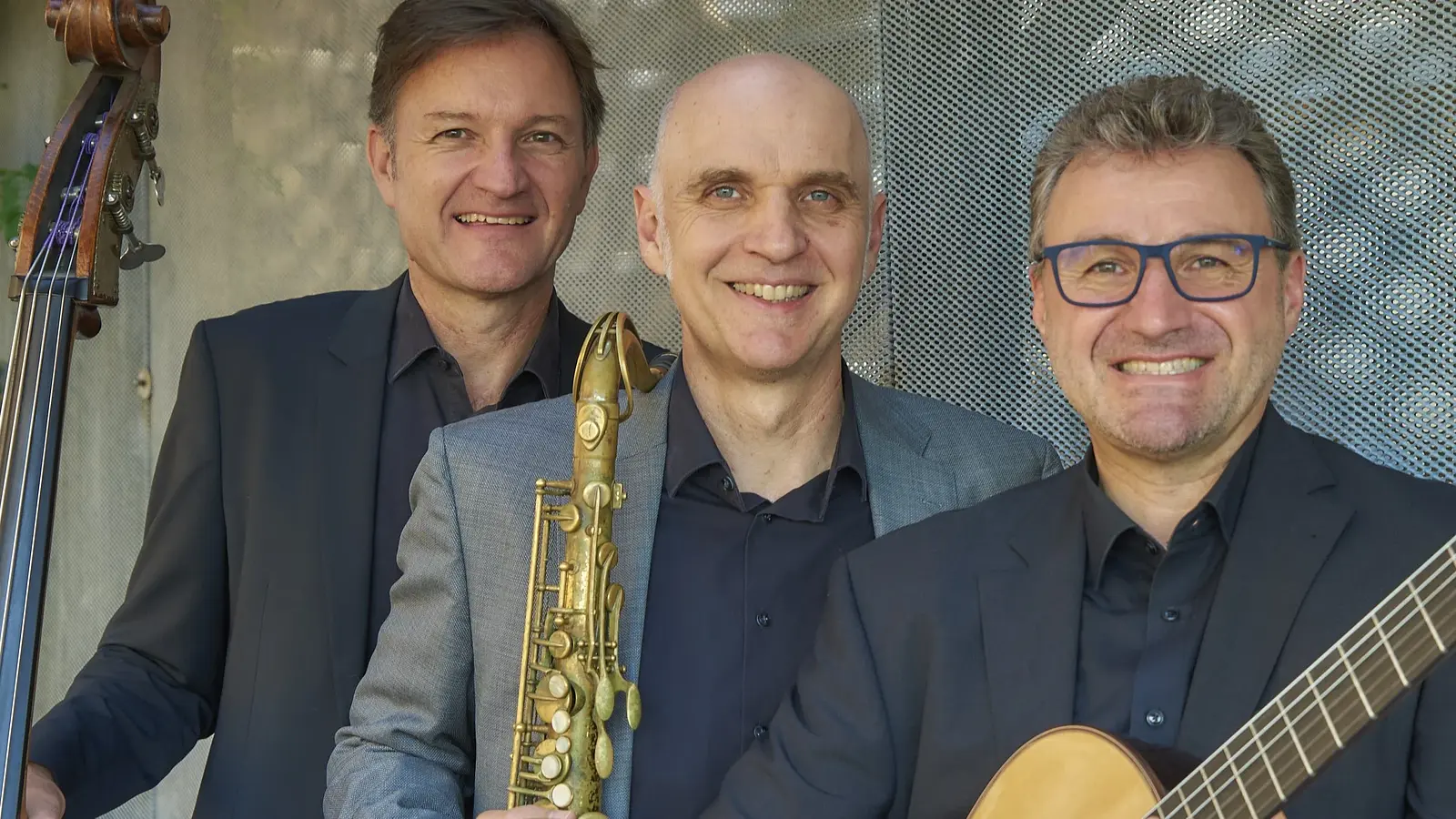Die drei Mitglieder der Golden Notes in Anzügen mit Instrumenten lächeln in die Kamera, vor verschwommenen grau-blauem Hintergrund