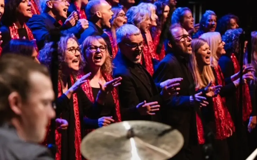 Gospelchor auf einer Bühne während eines Konzerts, im Vordergrund ein Schlagzeug, im Hintergrund singende Chormitglieder mit roten Schals