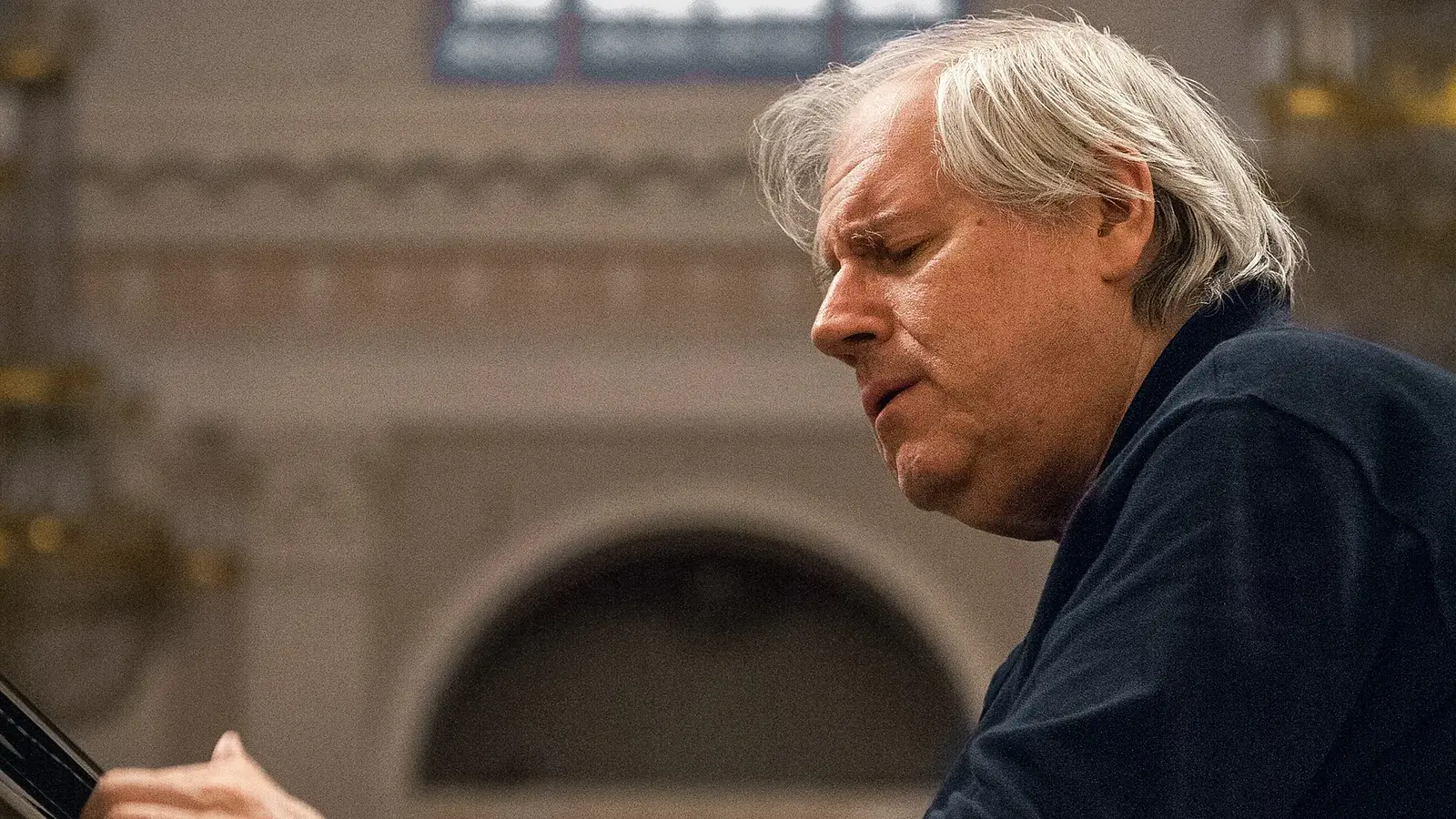 Promobild für das Event ‚Grigory Sokolov, Klavier‘: Eine Person in dunkler Kleidung sitzt an einem Flügel in einem großen Saal mit hellen Wandstrukturen und einem runden Bogen im Hintergrund