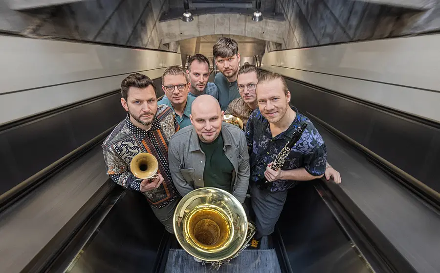 Sieben Musiker stehen mit Blas- und Holzblasinstrumenten auf einer Rolltreppe und blicken in die Kamera.