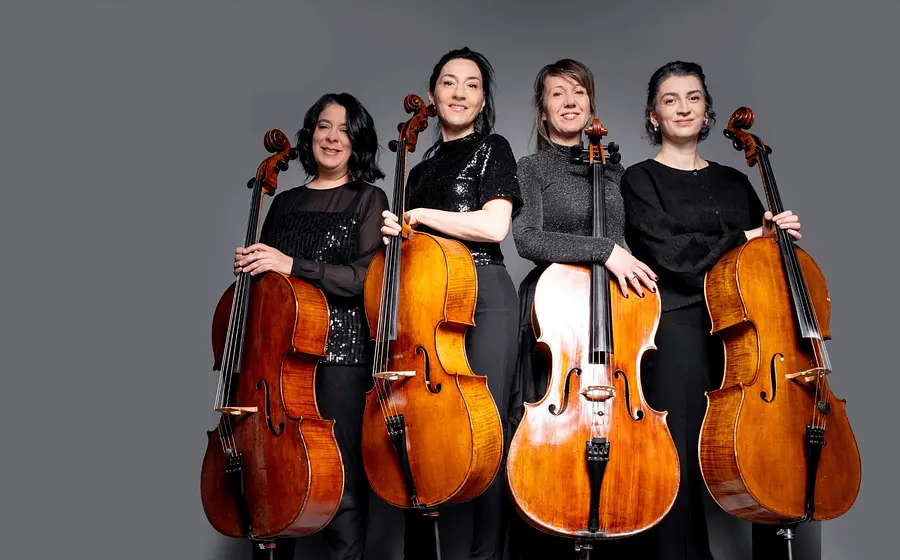 Gruppenporträt von vier Cellistinnen und Cellisten vor grauem Hintergrund. Die Musikerinnen und Musiker stehen nebeneinander in dunkler Konzertkleidung und halten ihre Violoncelli vor sich.